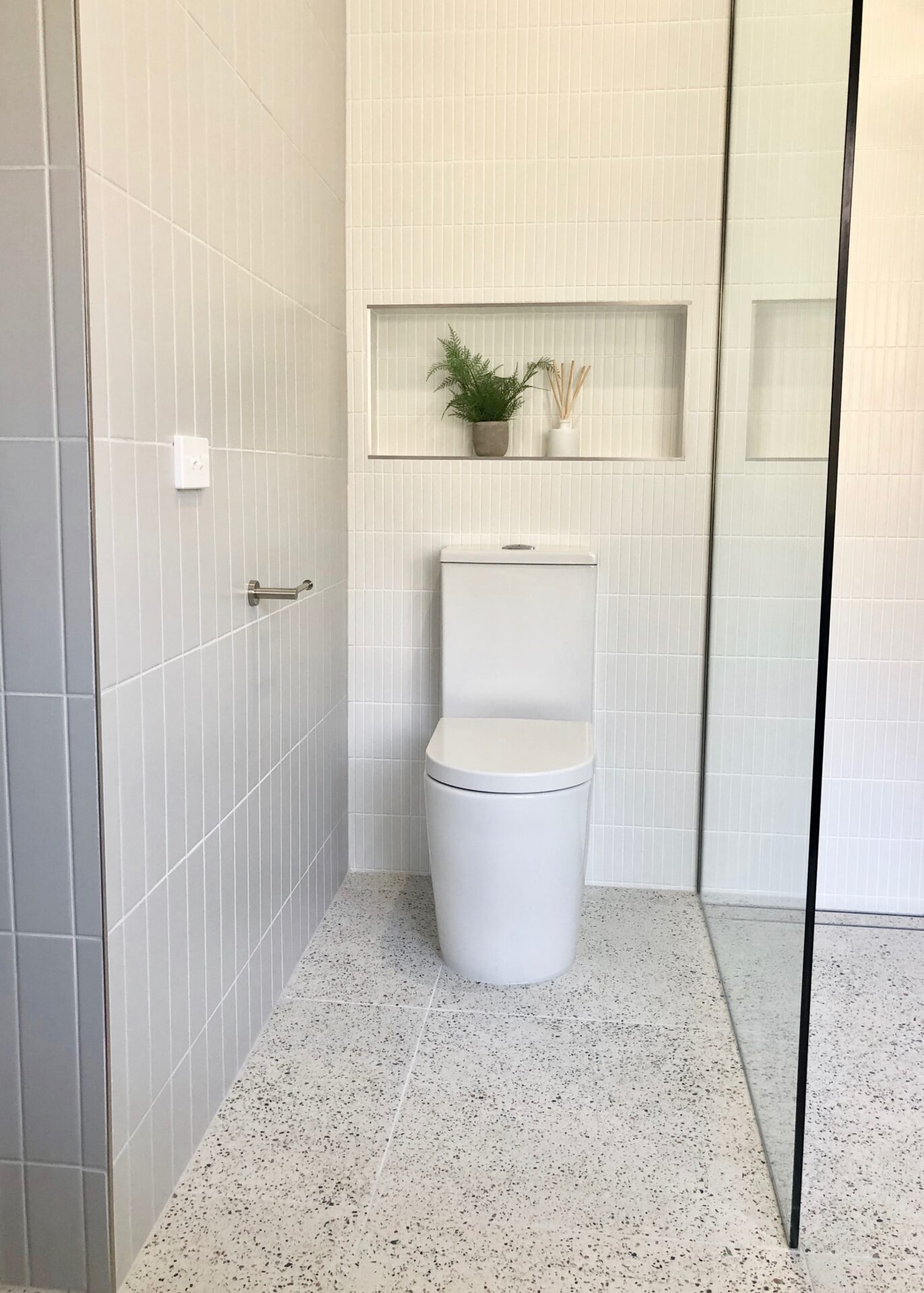 toilet in a white tiled bathroom with a plant vase and shower glass.